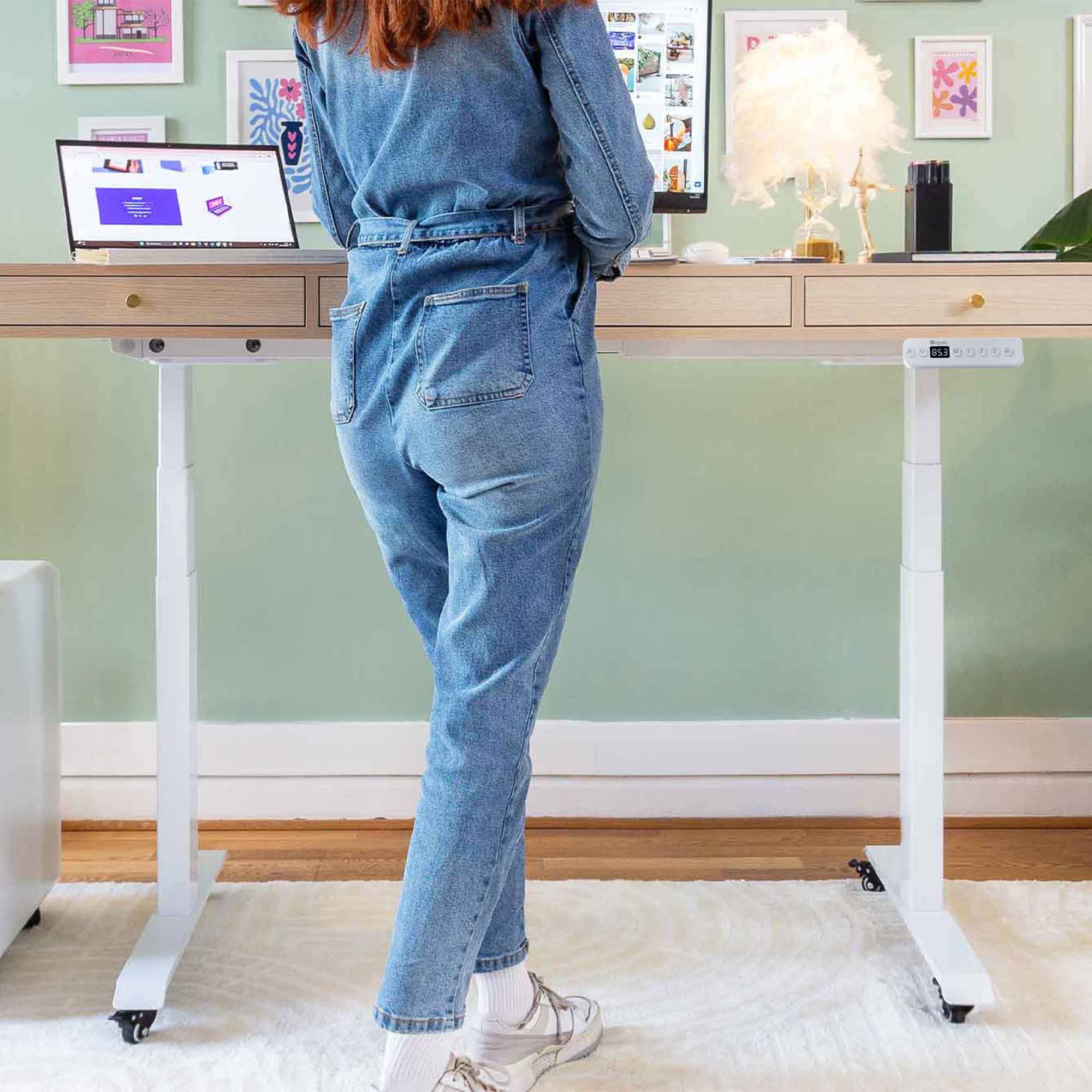 Bureau assis-debout KQUEO Space Dynamique en bois très clair avec 3 tiroirs et pieds blancs à roulettes, utilisé en position haute par une femme.